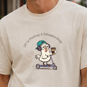 Puede incluir: Camiseta beige con un dibujo animado de un pollo en un scooter, con una gorra y auriculares color teal, y sosteniendo una taza de café. El texto "Off to Meetings & Debugging things" está arqueado sobre el pollo.