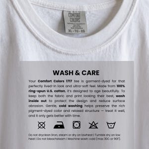Puede incluir: Una camiseta blanca Comfort Colors con una etiqueta en el cuello. La etiqueta dice "COMFORT COLORS XL TG EG". Se ve una hoja de instrucciones de cuidado, con texto sobre el lavado y las instrucciones de cuidado de la prenda.