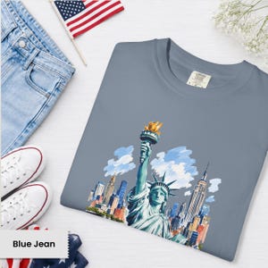 Puede incluir: Una camiseta azul gris&aacute;ceo con una ilustraci&oacute;n a la acuarela de la Estatua de la Libertad y el horizonte de la ciudad de Nueva York. La camiseta se muestra con vaqueros azules, zapatillas blancas y una peque&ntilde;a bandera americana.