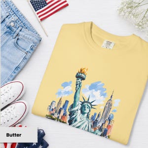 Puede incluir: Una camiseta de color mantequilla con una ilustraci&oacute;n en acuarela de la Estatua de la Libertad sosteniendo una antorcha, con el horizonte de la ciudad de Nueva York de fondo. La camiseta se combina con vaqueros azules, zapatillas blancas y una peque&ntilde;a bandera americana.