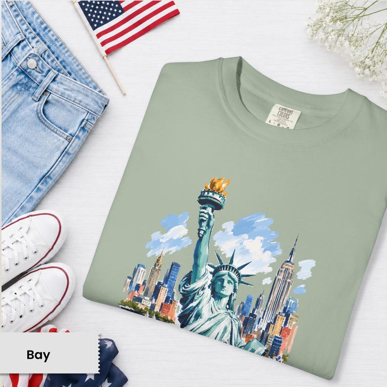 Puede incluir: Camiseta verde salvia con una ilustraci&oacute;n en acuarela de la Estatua de la Libertad y el horizonte de la ciudad de Nueva York. La camiseta incluye el logotipo de Comfort Colors en la etiqueta. La imagen est&aacute; combinada con vaqueros azules, zapatillas blancas y una bandera americana.
