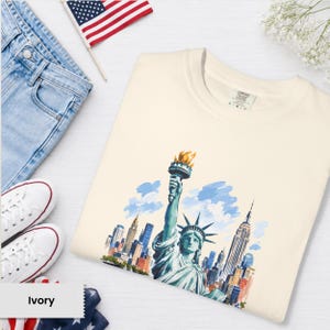 Puede incluir: Camiseta de color marfil con una ilustraci&oacute;n en acuarela de la Estatua de la Libertad y el horizonte de la ciudad de Nueva York. La camiseta se combina con vaqueros azules, zapatillas blancas y una peque&ntilde;a bandera americana.