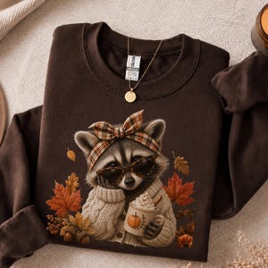 May include: A dark brown crewneck sweatshirt featuring a graphic of a raccoon wearing a plaid headband and sunglasses, dressed in a beige cable-knit sweater, holding a mug with a pumpkin design, surrounded by autumn leaves and acorns.