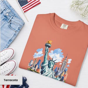 Puede incluir: Camiseta de color terracota con un gr&aacute;fico de la Estatua de la Libertad con el horizonte de la ciudad de Nueva York. La camiseta se muestra con vaqueros azules, zapatillas blancas y una peque&ntilde;a bandera americana. La camiseta tiene una etiqueta Comfort Colors.