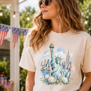 Puede incluir: Camiseta color crema con una ilustraci&oacute;n en acuarela de la Estatua de la Libertad y el horizonte de la ciudad de Nueva York. El dise&ntilde;o incluye detalles florales azules, verdes y amarillos. La camiseta es de un material suave.