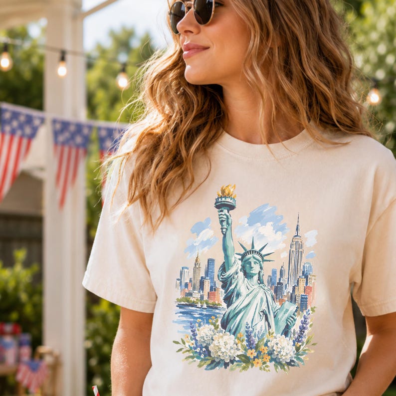 Puede incluir: Camiseta color crema con una ilustraci&oacute;n en acuarela de la Estatua de la Libertad y el horizonte de la ciudad de Nueva York. El dise&ntilde;o incluye detalles florales azules, verdes y amarillos. La camiseta es de un material suave.