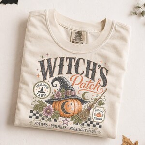 May include: A cream-colored t-shirt featuring a graphic print with the text "WITCH'S Patch". The design includes a pumpkin wearing a witch hat, a broomstick, flowers, and the text "SPELLBOUND FARM", "LOCALLY GROWN", "POTIONS PUMPKINS MOONLIGHT MAGIC".