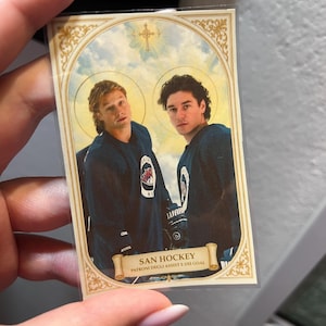 Könnte beinhalten: Eine Hockey-Sammelkarte mit zwei Männern in blauen Hockeytrikots. Das Design ist von religiösen Ikonen inspiriert, mit goldenen Akzenten und dem Text "SAN HOCKEY PATRONI DEGLI ASSISTI E DEI GOAL".