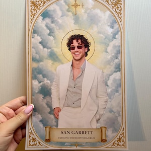 May include: A rectangular print featuring a smiling person with curly hair and sunglasses, wearing a light-coloured suit. The person is framed by a golden halo and clouds, with a scroll at the bottom reading SAN GARRETT PATRONO DEI BICIPITI DA URLO.