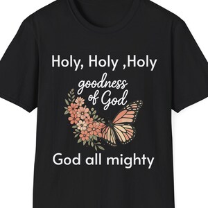 Puede incluir: Camiseta negra con el texto "Holy, Holy, Holy goodness of God God all mighty". Un gráfico floral y de mariposa está centrado debajo del texto. Las flores son melocotón y rosa, y la mariposa es naranja y marrón.