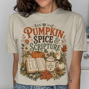 Może przedstawiać: Beżowy t-shirt z krótkim rękawem z nadrukiem graficznym i napisem PUMPKIN SPICE & SCRIPTURE. Wzór przedstawia otwartą książkę, kubek z bitą śmietaną, dynie, świece oraz jesienne liście w odcieniach pomarańczu, zieleni i brązu.