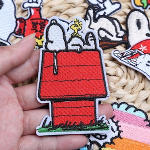 Puede incluir: Parche bordado con Snoopy y Woodstock encima de la caseta roja de Snoopy. El parche tiene un borde blanco y hierba verde en la base. Otros parches de personajes de dibujos animados son visibles en el fondo.