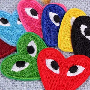 May include: A collection of colorful heart-shaped embroidered patches. The patches are in various colors, including blue, pink, green, yellow, red, and black. Each patch features a white eye with a black pupil.