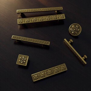 Puede incluir: Una colección de herrajes para muebles en tono latón antiguo que incluye cuatro tiradores rectangulares con patrones florales en relieve, un pomo cuadrado con un motivo floral central y un pomo circular con un diseño floral detallado.