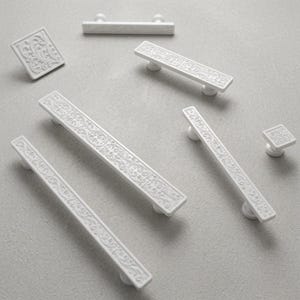 Puede incluir: Una colección de herrajes blancos para armarios que incluye cuatro tiradores rectangulares para cajones y dos pomos cuadrados. Cada pieza presenta un patrón floral en relieve intrincado en la superficie, proporcionando un acabado texturizado.