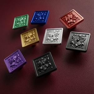 Könnte beinhalten: Eine Sammlung von acht quadratischen Schrankknöpfen in verschiedenen Farben, darunter Grün, Blau, Rot, Gold, Silber, Schwarz und Violett. Jeder Knopf zeigt ein erhabenes, geprägtes Blumenmuster in der Mitte mit detaillierten Blütenblättern.