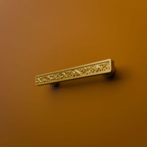 Könnte beinhalten: Ein rechteckiger Schubladengriff aus goldfarbenem Metall mit einem detaillierten, erhabenen Blumen- und Rankenrelief auf der Vorderseite. Der Beschlag ist horizontal montiert und weist eine polierte Oberfläche auf, die das Licht reflektiert.