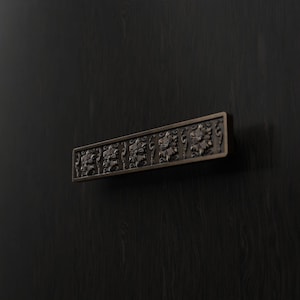 May include: A rectangular cabinet pull with a dark bronze finish, featuring a repeating embossed floral pattern across its front surface. The hardware is mounted horizontally, showcasing intricate carved details and a textured metal construction.