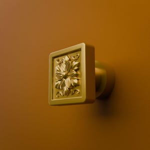May include: A square gold-toned cabinet knob featuring a raised floral relief pattern in the center. The knob has a smooth, matte finish and a cylindrical base that extends outward from the square face, mounted on a solid mustard-colored surface.