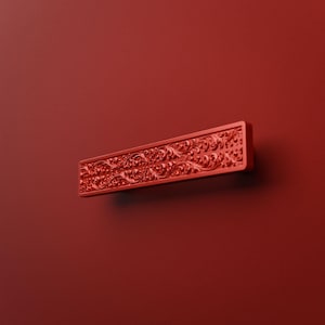 Peut inclure: Une poignée de meuble rectangulaire dans une finition rouge mat unie. La surface de la poignée présente un motif complexe de volutes florales en relief qui s'étend sur toute la longueur de la pièce, offrant un aspect texturé et détaillé.