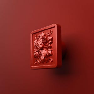 May include: A square-shaped cabinet knob in a solid matte red finish. The front face features a raised, embossed floral design with intricate petal details and small scrollwork accents at the top and bottom corners of the square frame.