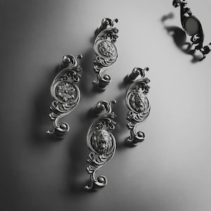 Könnte beinhalten: Eine Sammlung von vier dunkelgrauen Metall-Schubladengriffen mit komplexen Schnörkeln und Reliefdesigns. Zwei Griffe zeigen ein Löwenkopfmotiv, einer ein florales Muster und einer ein kreisförmiges Filigrandesign mit einer glatten Mitte.