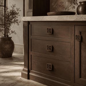Peut inclure: Un meuble en bois brun foncé doté de trois tiroirs superposés et d'une porte latérale, chacun équipé d'un bouton carré présentant un motif floral sculpté en relief. Le meuble possède une finition mate lisse et des détails cannelés verticaux.