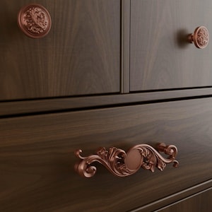 May include: A dark brown wooden cabinet featuring two round knobs and one horizontal handle, all finished in a metallic copper tone. The hardware displays intricate, raised floral scrollwork patterns, contrasting against the smooth wood grain.