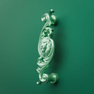 May include: A vertical cabinet handle in a matte mint green finish featuring an ornate, raised relief design of a lion head centered within swirling acanthus leaf scrollwork. The hardware is mounted to a matching solid mint green surface.
