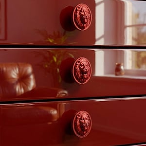 Pode incluir: Três puxadores de gaveta circulares com acabamento em vermelho profundo, cada um apresentando um relevo detalhado de cabeça de leão em três dimensões. Os puxadores estão montados numa superfície de armário vermelho escuro de alto brilho.