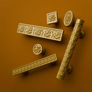 Puede incluir: Una colección de herrajes dorados para armarios que incluye dos tiradores rectangulares con patrones florales en relieve, un pomo circular con centro floral, un pomo cuadrado, un pomo ovalado y un tirador cilíndrico largo con superficie texturizada.