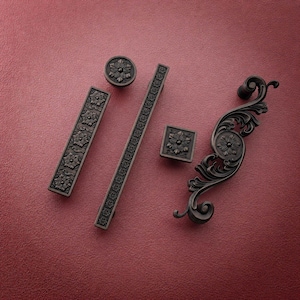 Op de afbeelding: Een collectie kastbeslag van donker brons, inclusief een ronde knop, een rechthoekige greep, een lange lineaire greep, een vierkante knop en een gebogen, sierlijke handgreep. Elk stuk is voorzien van een verhoogd bloemmotief.