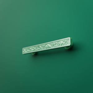 May include: A rectangular drawer pull handle featuring an intricate, raised floral scrollwork pattern across its front surface. The hardware is finished in a light, pale green color and is mounted horizontally against a solid dark green background.