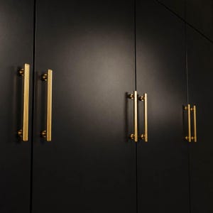 以下が含まれることがあります： マットブラックのキャビネットドアに、垂直で長方形のゴールドトーンの金属製ハンドルが取り付けられています。各ハンドルは2つの円形マウントで固定されており、暗く滑らかなキャビネットパネルの表面に対して、際立ったコントラストを生み出しています。