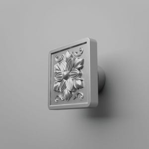 Op de afbeelding: Een vierkante zilverkleurige meubelknop met een verhoogd bloemmotief in het midden. Het ontwerp bevat gedetailleerde bloemblaadjes en kleine krulaccenten binnen een gladde, verhoogde rand, bevestigd aan een cilindrische basis.