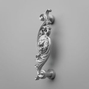 May include: A silver-toned metal cabinet handle featuring an ornate, swirling scrollwork design. The center of the handle displays a detailed, raised relief of a lion's head facing forward. The handle is mounted on two cylindrical posts.