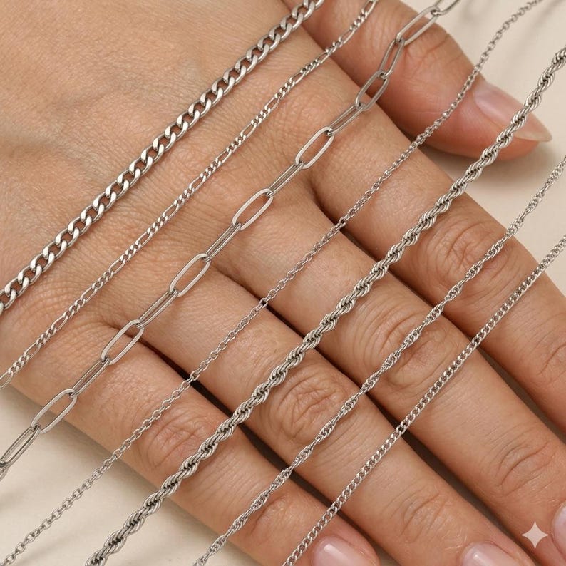 May include: A collection of silver necklaces with various chain styles, including curb, oval link, rope, and cable chains. The necklaces are displayed on a hand, showcasing the different designs and textures of the jewelry.
