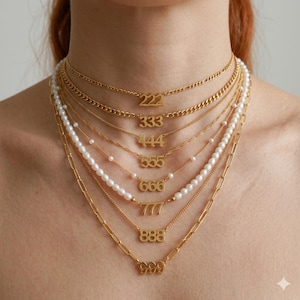 May include: A collection of gold-colored necklaces with various number pendants, including 222, 333, 444, 555, 666, 777, 888, and 999. Some necklaces feature delicate pearl strands, while others have chain links. The necklaces are layered for a stylish effect.