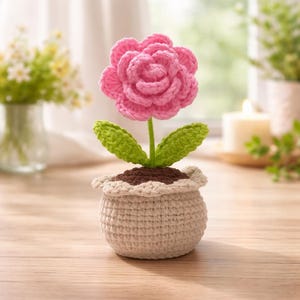 Pode incluir: Uma flor de crochê feita à mão em um vaso. A flor rosa tem várias camadas de pétalas, folhas verdes e um caule verde. O vaso é bege com uma borda com babados e preenchido com terra marrom.