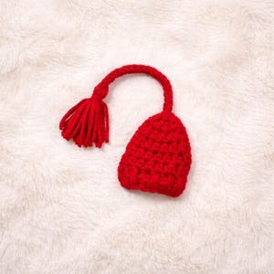 Pode incluir: Um item vermelho de crochê feito à mão com uma borla e uma forma arredondada, semelhante a um chapéu. O item é feito de um fio macio e texturizado e é colocado em um fundo branco e fofo. A borla pende de um caule curvo.