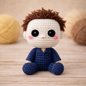 May include: A handmade crochet doll of a character with a white head, brown hair, and a navy blue jumpsuit. The doll has large black eyes, pink cheeks, and is sitting. The background includes balls of yarn.