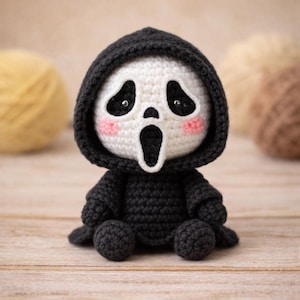 Peut inclure: Poupée Ghostface au crochet faite à la main, représentant le personnage emblématique du film Scream. La poupée a un visage blanc avec des orbites noires et une bouche ouverte, portant une robe à capuche noire. Les joues sont roses. La poupée est assise sur une surface en bois.