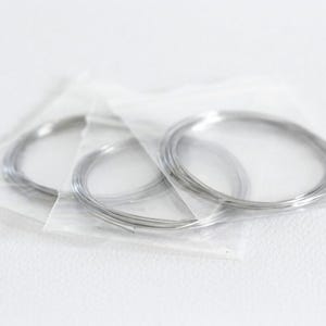 Könnte beinhalten: Drei separate Spulen aus dünnem, silberfarbenem Metalldraht, die jeweils einzeln in einem kleinen, durchsichtigen, rechteckigen Plastikbeutel verpackt sind. Der Draht ist zu kreisförmigen Schlaufen gewickelt und weist eine glatte, reflektierende Oberfläche auf.