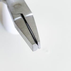 Può includere: Una pinza in metallo con ganasce lunghe e affusolate, caratterizzate da una superficie interna piatta e un profilo esterno arrotondato. L'utensile ha una finitura metallica argentata ed è dotato di impugnature in gomma blu scuro.