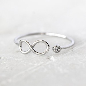 Op de afbeelding: Een zilverkleurige open ring met een oneindigheidssymbool aan het ene uiteinde en een enkele kleine, heldere, rondgeslepen steen in een ronde metalen zetting aan het andere uiteinde. De ringband is dun en gepolijst voor een strakke uitstraling.