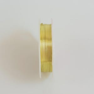 Puede incluir: Una bobina de alambre fino de color oro metálico enrollado firmemente alrededor de un núcleo circular de plástico blanco. El alambre está dispuesto en capas paralelas y uniformes, creando una superficie lisa y reflectante en todo el ancho.