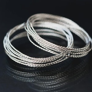 Pode incluir: Um conjunto de várias pulseiras finas de metal prateado com uma superfície torcida e texturizada. As pulseiras estão dispostas numa formação circular sobreposta, criando uma aparência em camadas com um acabamento metálico reflexivo.