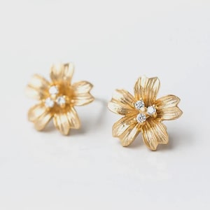 Peut inclure: Une paire de boucles d'oreilles à tige en forme de fleurs à cinq pétales. Chaque boucle présente une finition dorée métallique texturée sur les pétales et trois petites pierres rondes et transparentes regroupées au centre du motif floral.