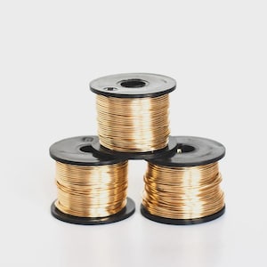 Puede incluir: Tres bobinas de plástico negro apiladas en forma de pirámide, cada una enrollada firmemente con una longitud continua de alambre metálico de color dorado. Las bobinas presentan un diseño circular con un orificio central, mostrando el alambre.