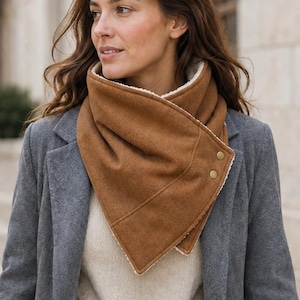 May include: A brown triangular scarf with a white fleece lining and two gold-toned metal snap buttons. The scarf is worn over a beige sweater and a long grey wool coat. The design features a pointed front and a wrap-around style neck closure.
